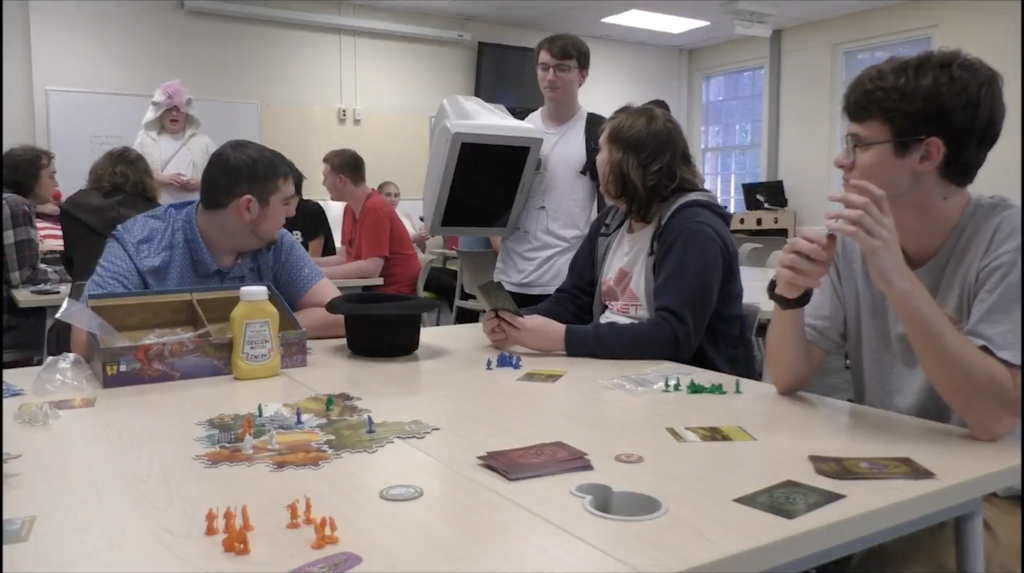 A group of students sits around a large table covered in cards, miniature figures, and other games pieces as they talk about the board game they are playing.