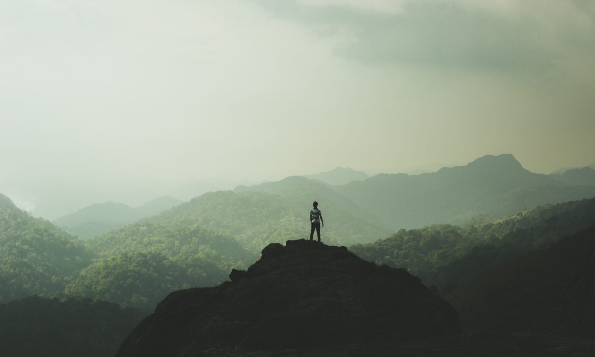 wide angle photo of a man standing on a cliff looking out at mountains in the distance