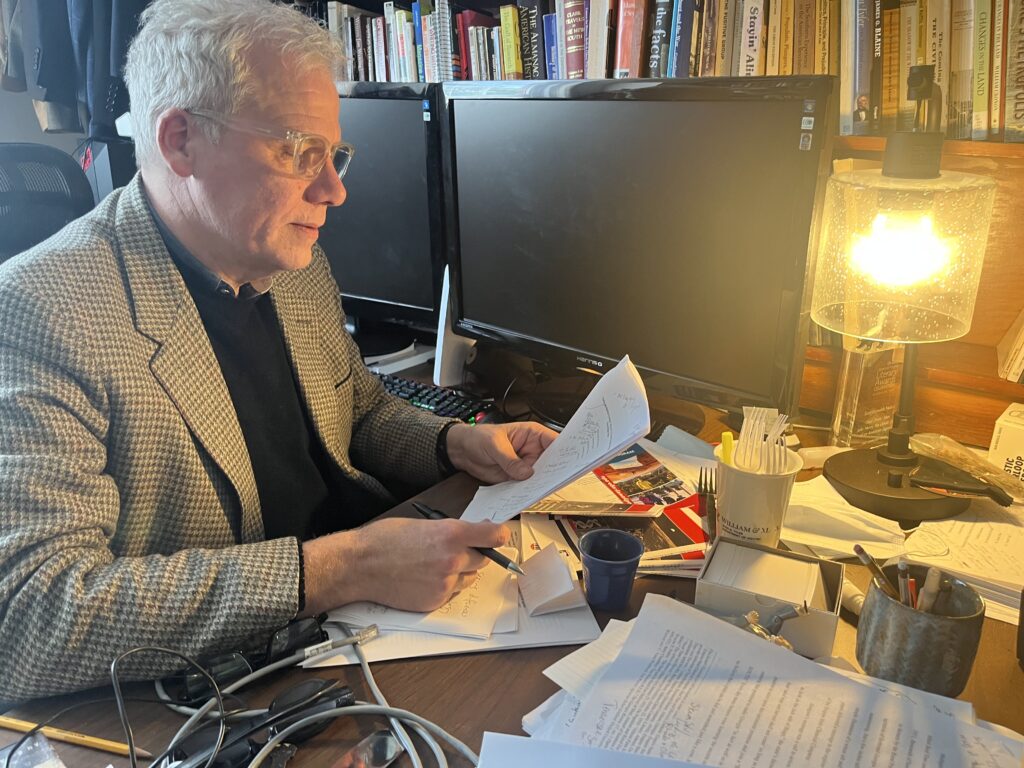 Scott Reynolds Nelson reads an essay at a desk covered in similar essays while a lamp lights up his face and the bookshelf behind him